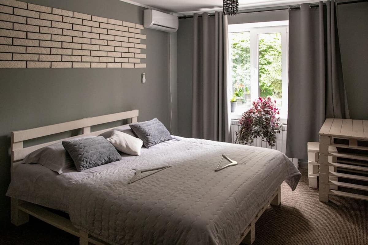 A bedroom with a bed, a brick wall, an air conditioning unit, a window with curtains, and a plant.
