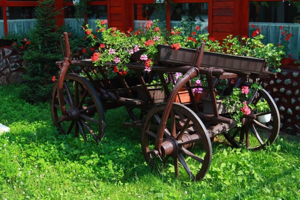 A wooden cart with flower boxes, surrounded by greenery and a red building in the background.