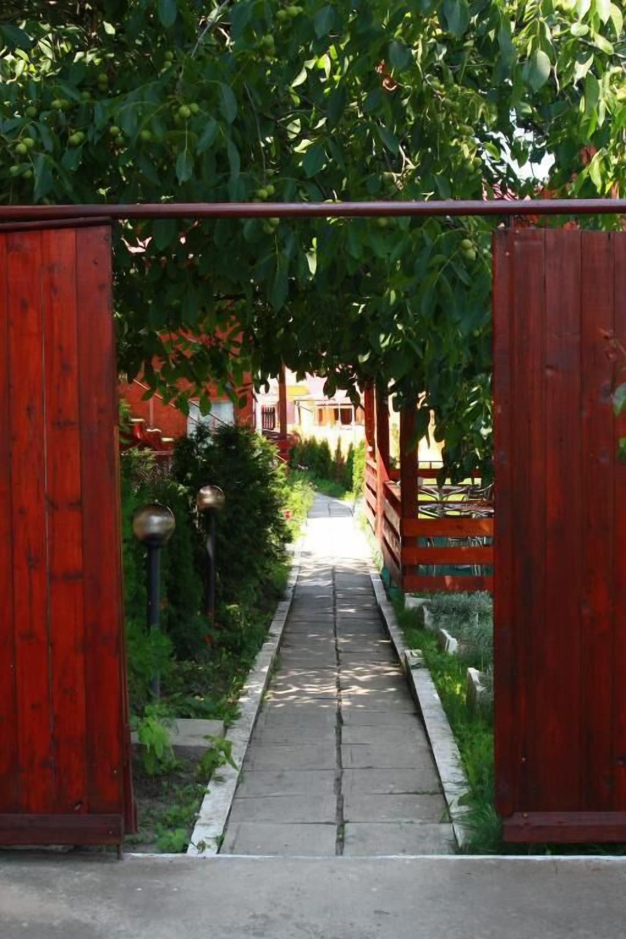 A wooden gate leading to a pathway lined with greenery and a building in the background.