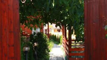 A wooden gate leading to a pathway lined with greenery and a building in the background.