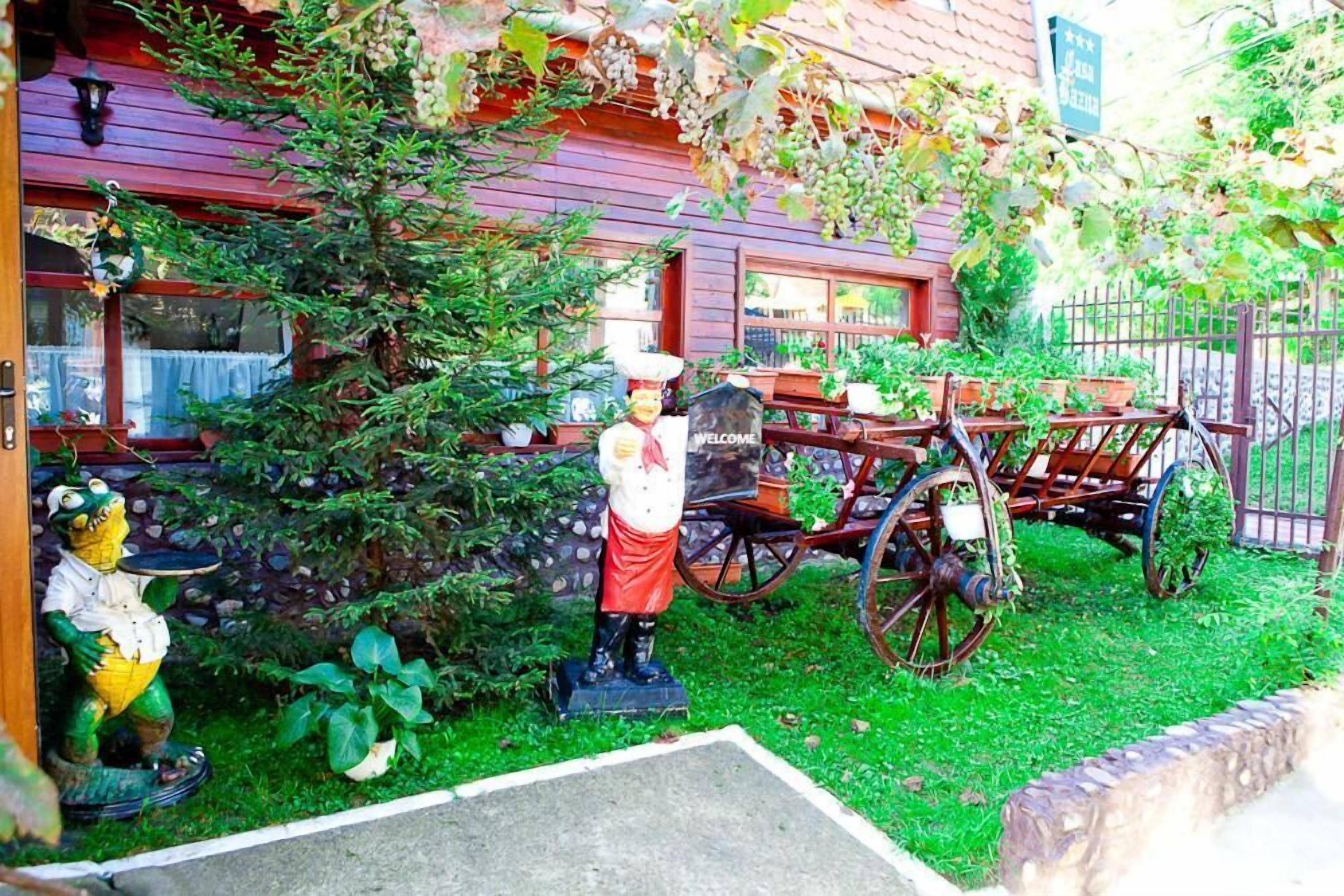 Un edificio rústico con fachada de madera, un letrero de bienvenida, una estatua de oso, un carrito antiguo y un pequeño jardín.