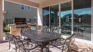 Eine Terrasse im Freien mit einem runden Metalltisch und Stühlen, einem Grill und einer Glastür, die zu einem Poolbereich führt.