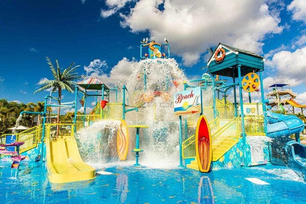 A water park with multiple slides and water features, including a large water fountain and a sign indicating a beach area.