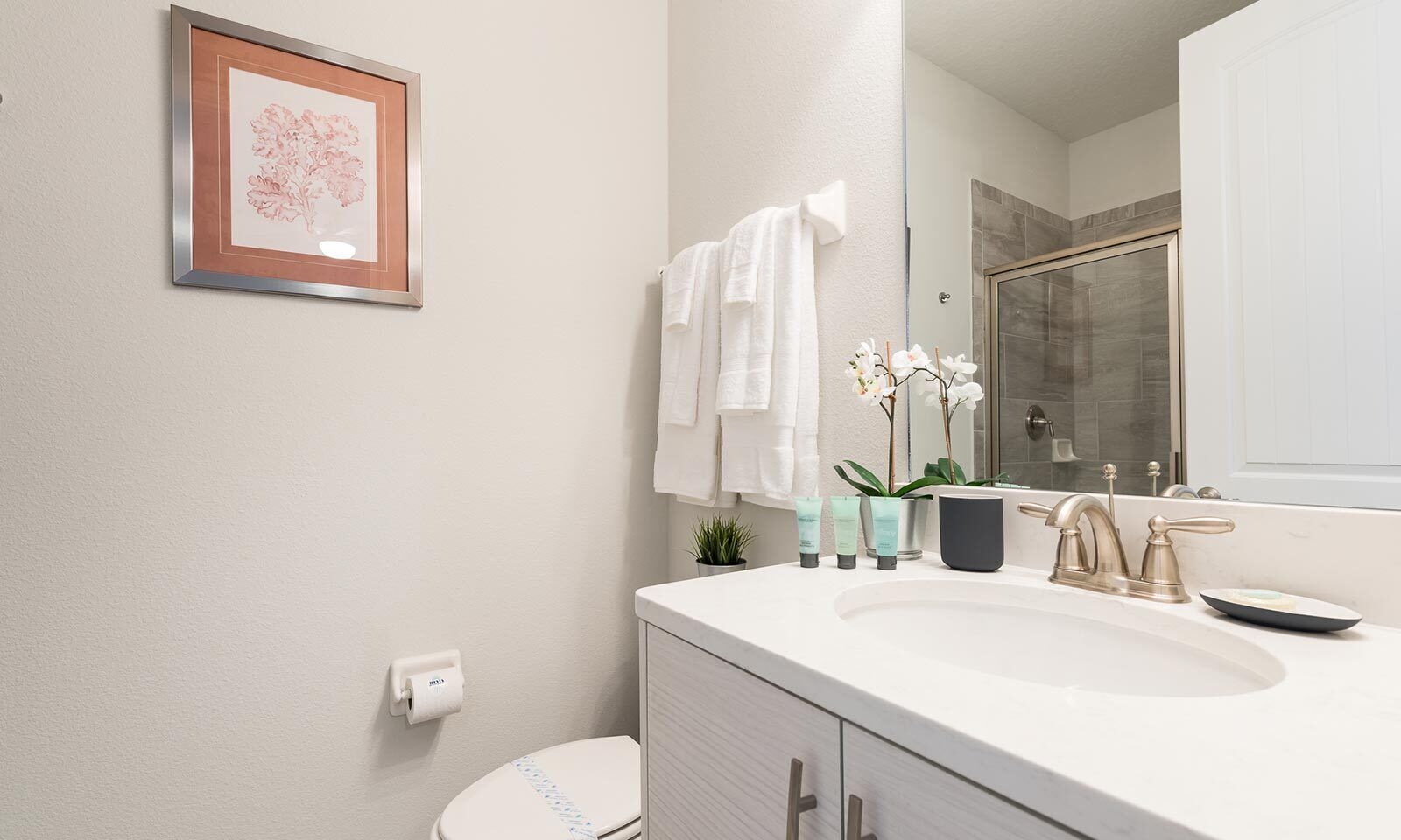 A modern bathroom with a white sink, two faucets, a framed picture on the wall, a toilet, and a shower area visible through a glass door.