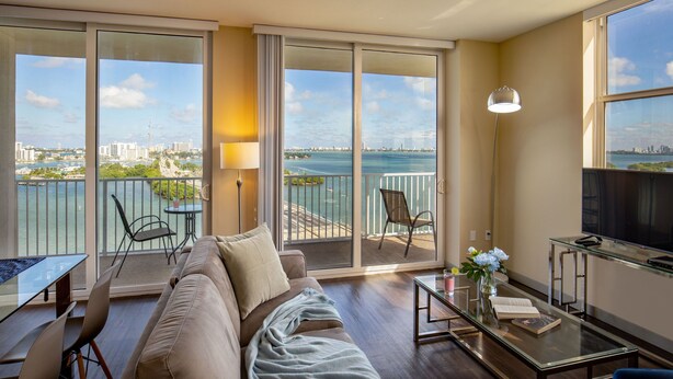 A modern living room with a sofa, a glass coffee table, and a TV. There is a balcony with a view of the city and a body of water. - Shorecrest Miami Bay (Miami)