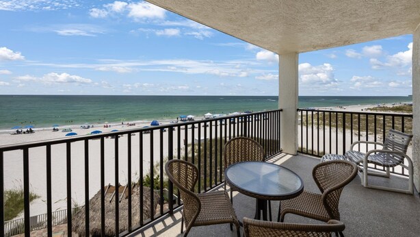 Un balcon avec des fauteuils en osier et une table, donnant sur une plage parsemée de parasols et fréquentée par des gens.