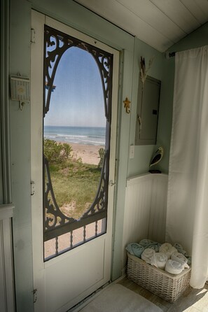 Vista da praia através de uma porta decorativa, um cesto de vime com toalhas e uma decoração de pássaro na parede.