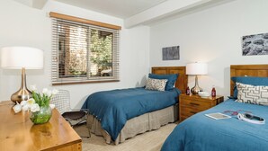 Un dormitorio con dos camas, un aparador de madera, un escritorio, una silla, un ventanal con persianas y un cuadro en la pared.