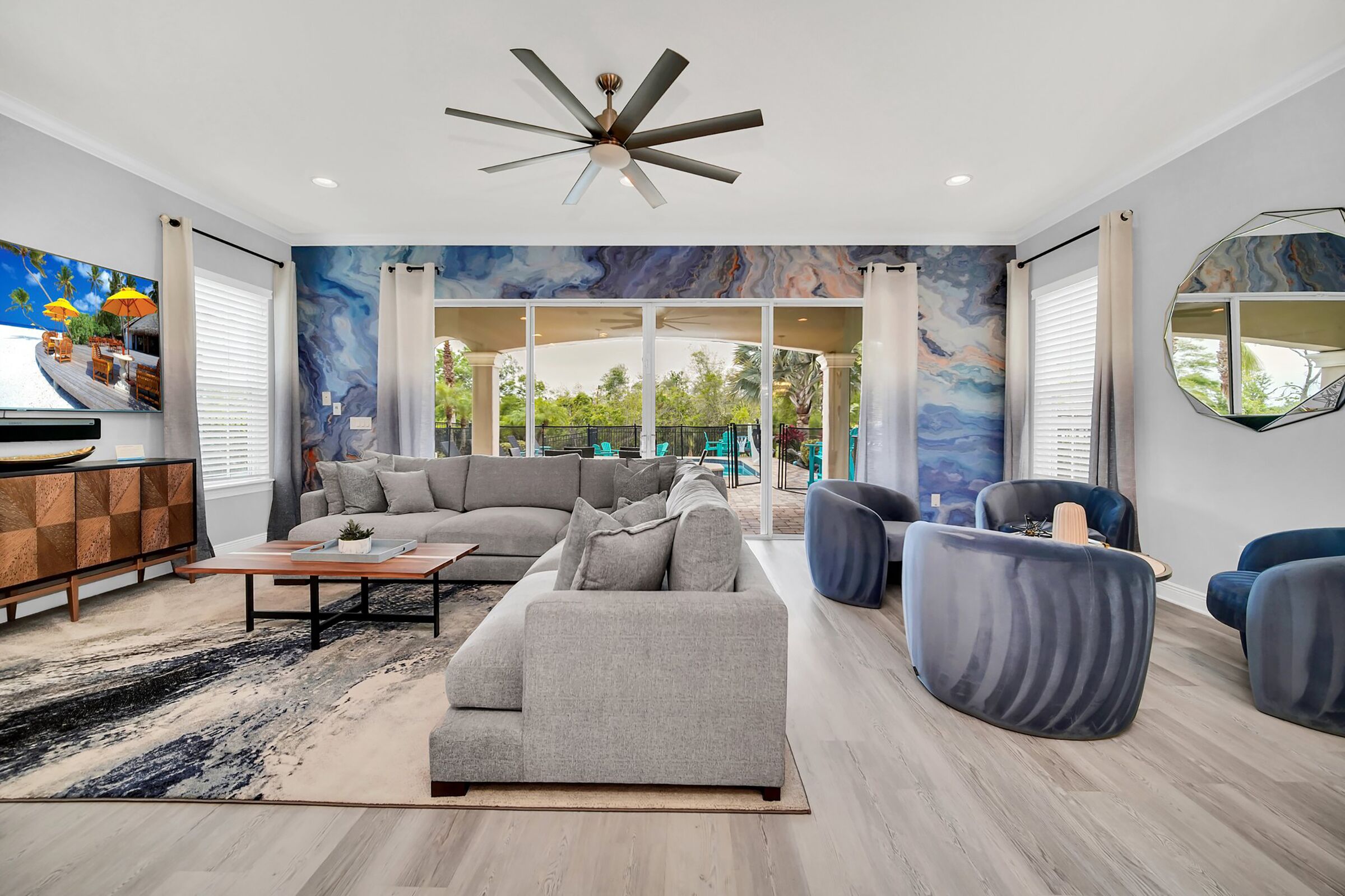A modern living room with a large sectional sofa, a coffee table, and a TV on a wooden cabinet. There is a ceiling fan, a large window, and a view of the outdoors.