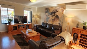A living room with a leather sofa, wooden coffee table, and a TV on a wooden cabinet. There is a large window with a view outside, a wall-mounted air conditioner, and a bookshelf with books and decorative items. - Palm Beach Manor ... (Rockingham)