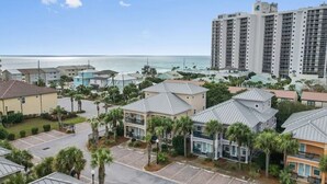 A coastal residential area with a mix of single and multi-story buildings, some with balconies, and a parking lot in the foreground. - Spacious Rental, Three Story Townhome, Close to Pool (Miramar Beach)