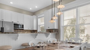 A modern kitchen with white cabinets, a wooden dining table, and pendant lights. - Spacious Rental, Three Story Townhome, Close to Pool (Miramar Beach)