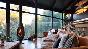 A modern living room with a large fireplace, a leather sofa, and a view of trees through large windows.