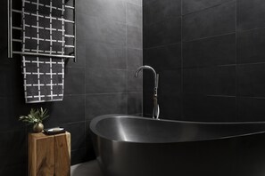 Un baño moderno con una bañera independiente, una mesita de madera con un florero y un perchero con una toalla de cuadros.