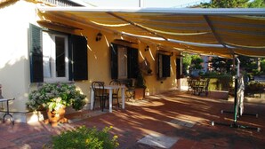 Alpendre coberto com mesa, cadeiras, plantas em vasos e um toldo amarelo.