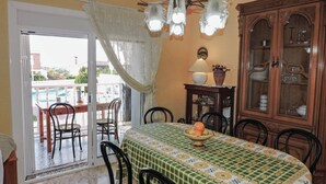 Un coin repas avec une table recouverte d’une nappe à motifs, entourée de chaises. On aperçoit, sur la droite, un meuble avec des portes en verre.