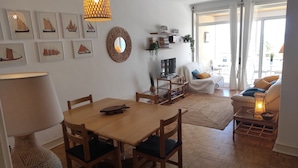 Een woonkamer met een houten eettafel, stoelen, een televisie op een dressoir, een bank en een bijzettafeltje. Er hangen ingelijste foto's aan de muur, er staat een geweven mand en een potplant.
