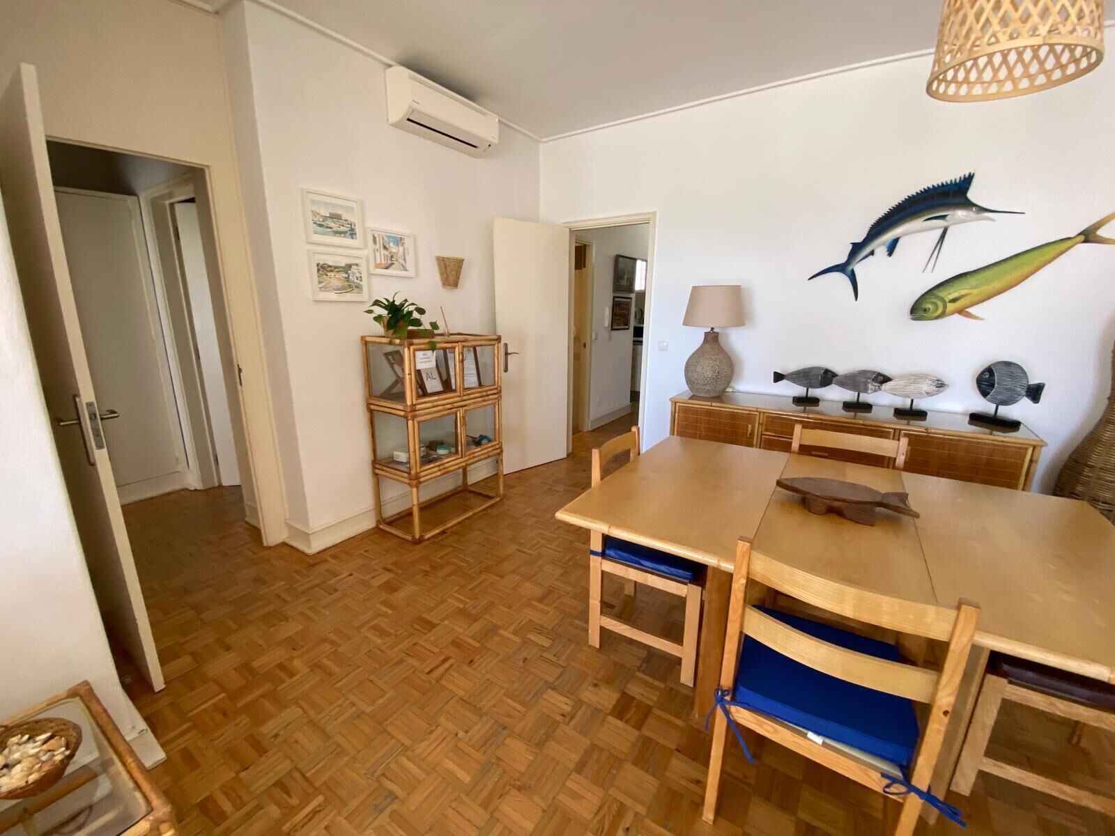 Un comedor con una mesa y sillas de madera, una mesita auxiliar de mimbre con una lámpara, y decoraciones de peces montadas en la pared.