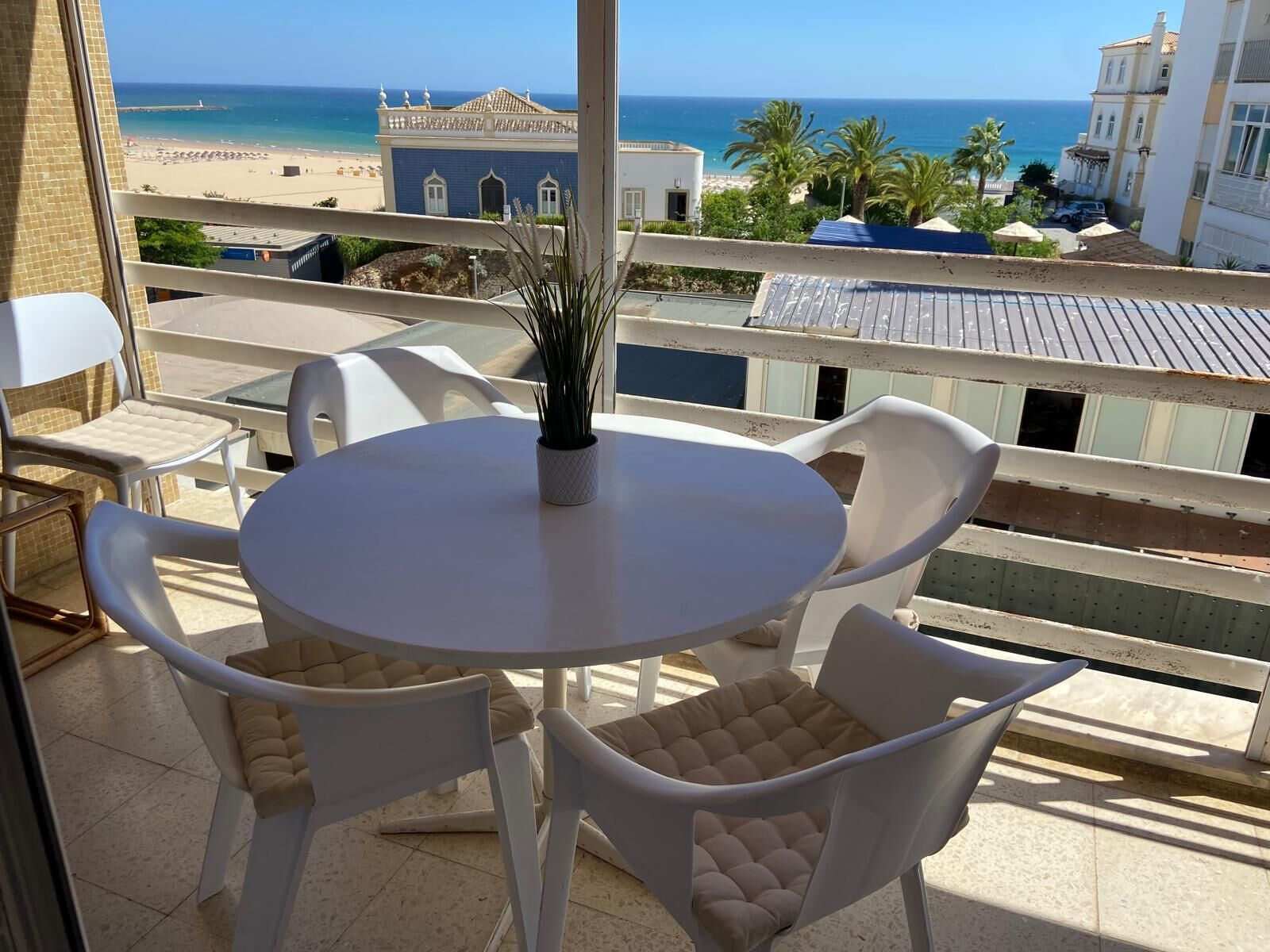A balcony with a round table and four chairs, overlooking a beach and buildings.