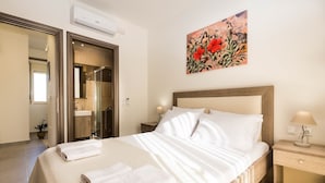 Une chambre d’hôtel comprenant un lit, une table de chevet, un climatiseur fixé au mur, une salle de bain avec un miroir et un tableau représentant des fleurs.