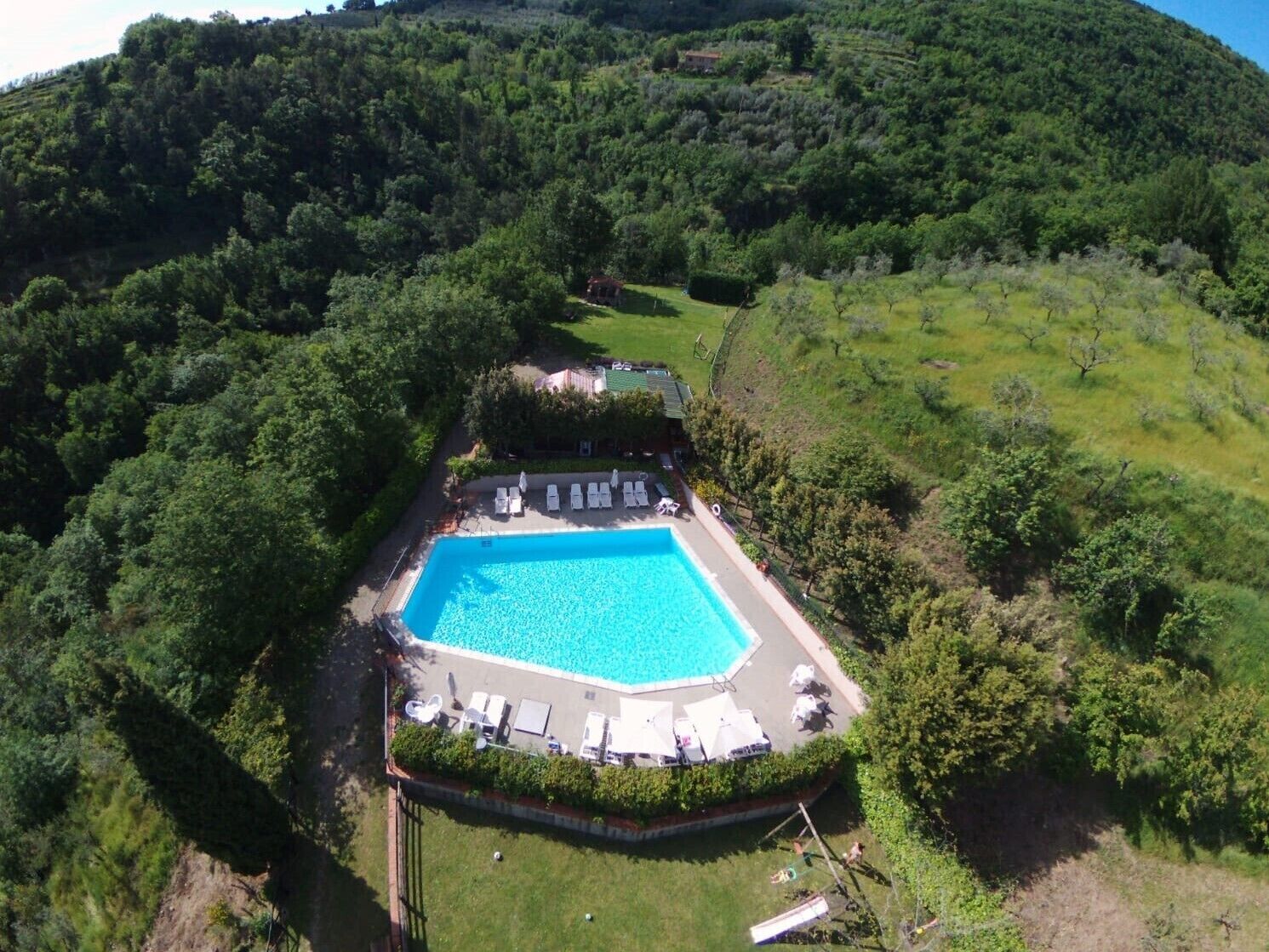 Une vue aérienne d’un complexe hôtelier avec une piscine rectangulaire entourée de chaises longues et de parasols, le tout se détachant sur un paysage verdoyant et vallonné.