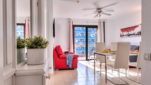 A modern living room with a red sofa, a dining table with chairs, a ceiling fan, and a balcony with a view of a building. - LOVELY 2 BEDROOMED APARTMENT NEXT TO THE BEACH (Arona)