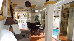 A sunlit room with a sofa, a table, chairs, and a view of a swimming pool through glass doors. - Town House With Private Pool ,Good WiFi. Short walk to Villamartin Plaza (alicante)