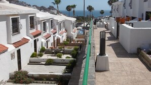 A coastal street with white buildings, palm trees, and a view of the sea. - "🏖 Double terrace with sun all day - 2 bedrooms - Perfect for 4 - Mogán (Mogán)