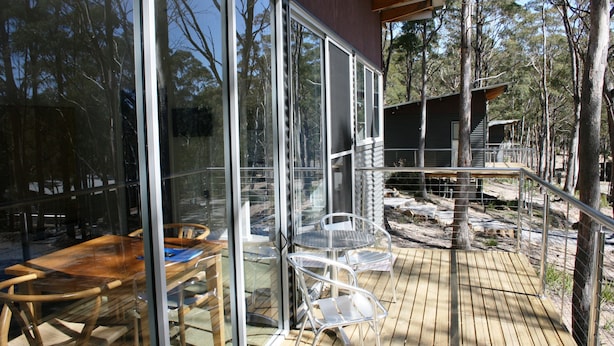 Terraza de madera con sillas y una mesa de metal, rodeada de paredes de vidrio que ofrecen vistas a árboles y un edificio.