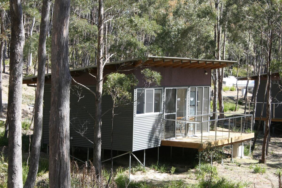 A small cabin with a wooden deck and a large window, surrounded by trees.