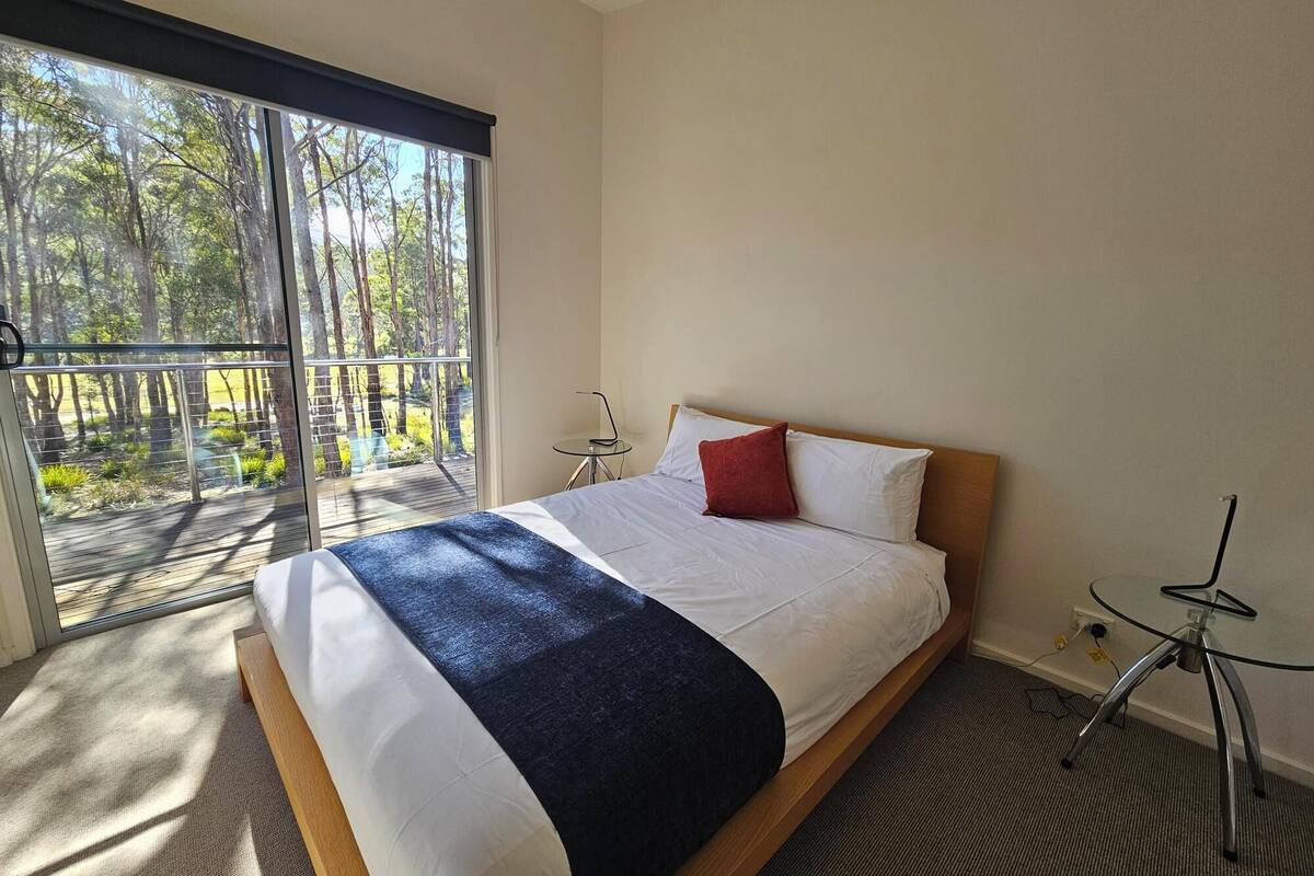 A bedroom with a large bed, a small bedside table, a glass table, and a view of a forest through a sliding door.