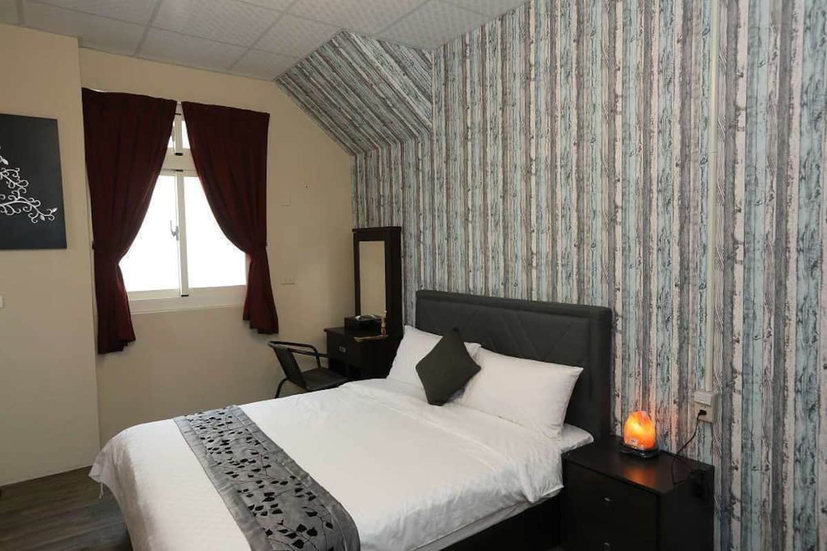 A neatly made bed with a headboard, a bedside table with a lamp, and a window with curtains.
