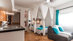 Een compacte hotelkamer met een kitchenette, een bed met hemelbed, een bank en een groot raam met gordijnen.