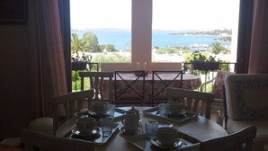 Ein Esstisch ist gedeckt mit Teetassen und Untertassen, einem Glas und einer Teekanne, mit Blick auf das Meer und die grüne Landschaft.