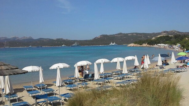 Ein Strand mit Reihen von Sonnenschirmen und Liegestühlen, einem klaren blauen Meer und Booten in der Ferne.