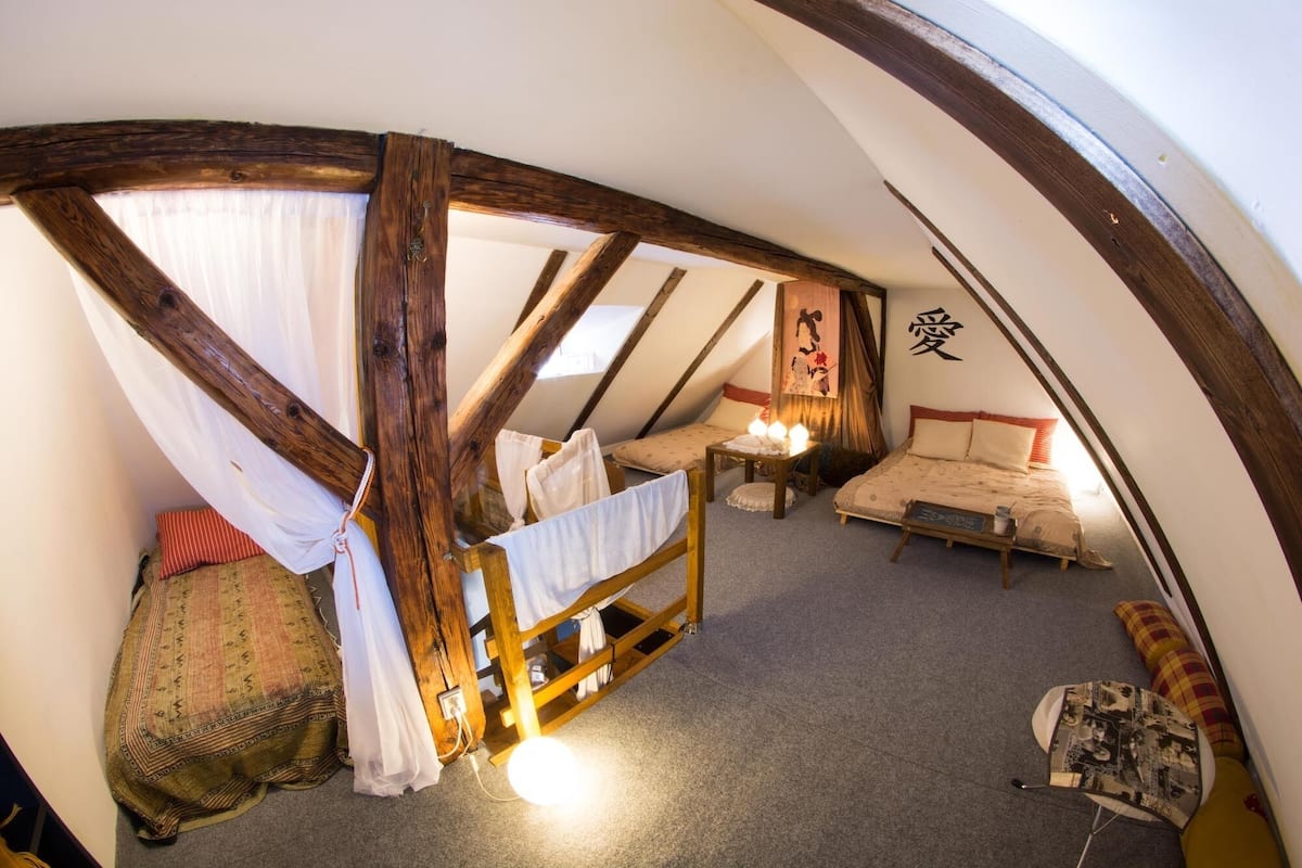 A cozy attic bedroom with a sloped ceiling, wooden beams, a bed, a small table, and a chair.