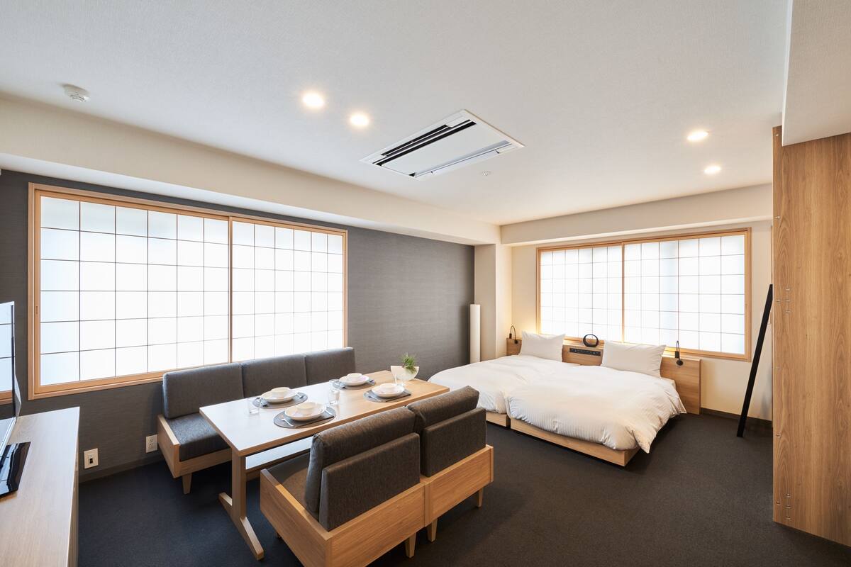 A modern hotel room with a bed, a dining table with chairs, a sofa, and large windows.