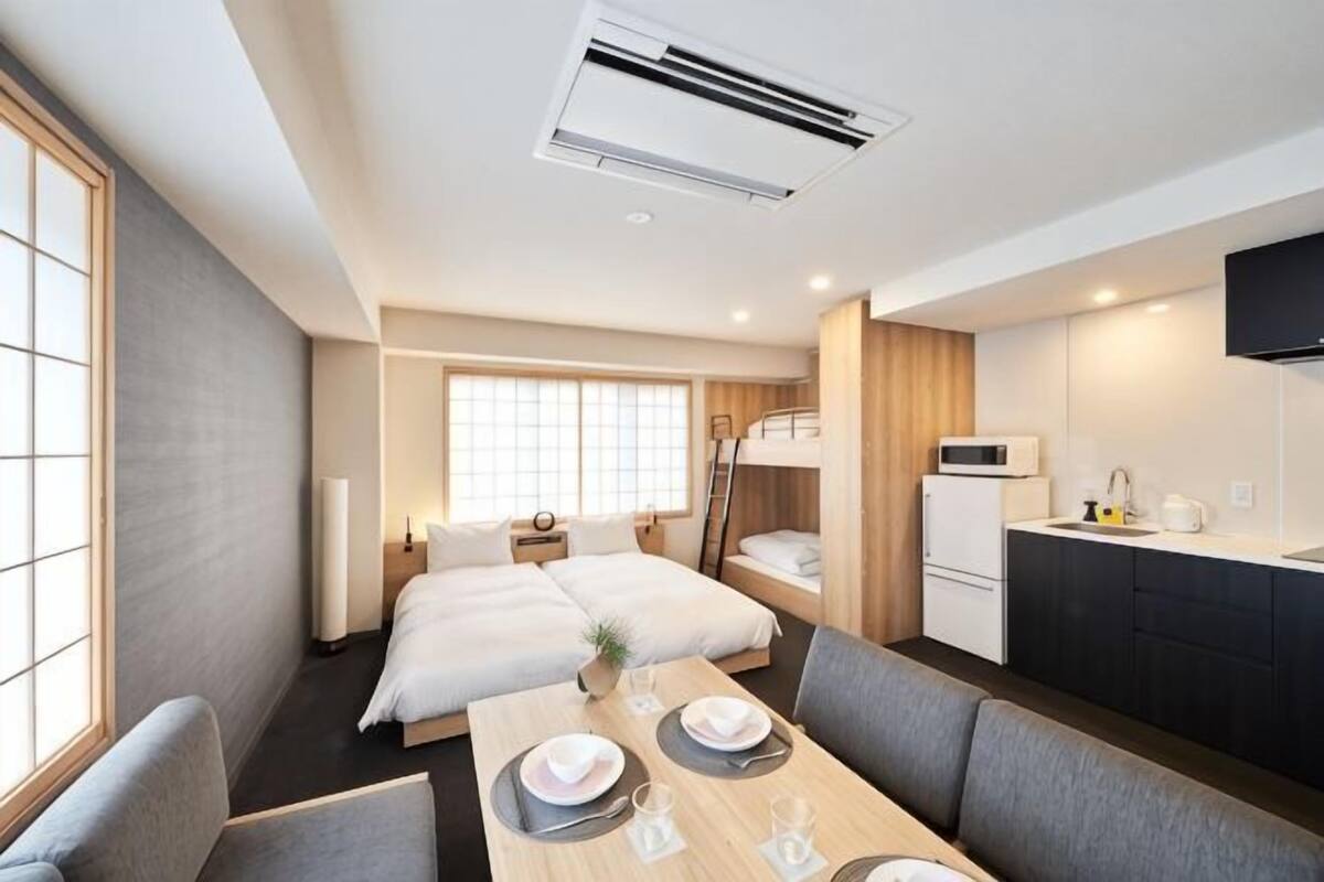 A hotel room with a bed, a dining table with chairs, a kitchen area, and a TV mounted on the wall.