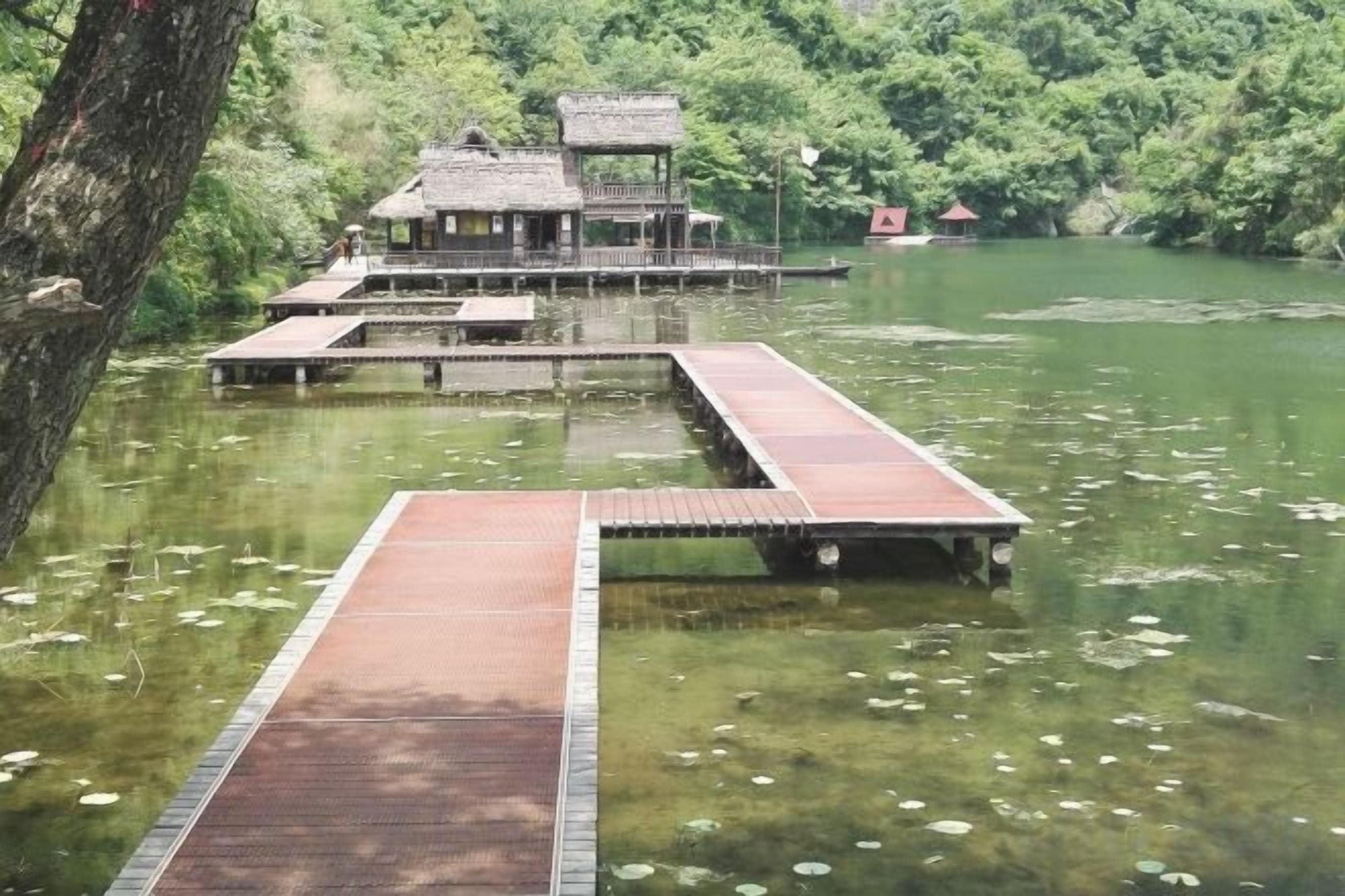 A wooden dock leading to a traditional building with a thatched roof, surrounded by a calm lake with lily pads.