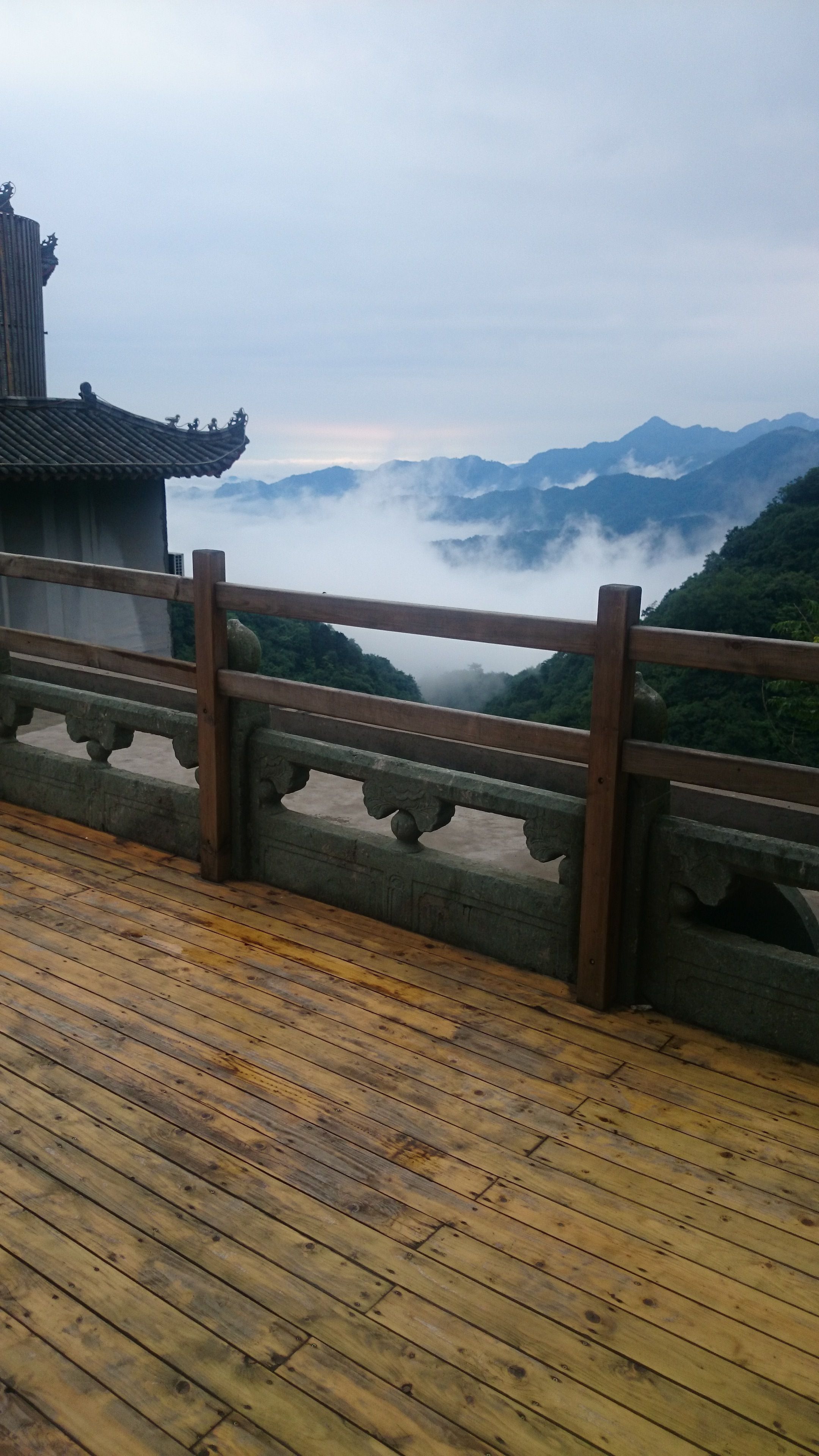 Una terraza de madera con barandilla que da a un paisaje montañoso cubierto de niebla y arquitectura tradicional.