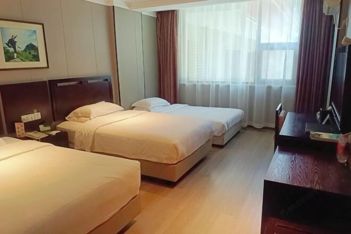 A hotel room with two beds, a wooden desk, a TV, and a window with sheer curtains.