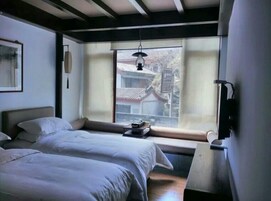Quarto de hotel com duas camas, janela com vista para um edifício tradicional, luminária pendente e TV fixada na parede.
