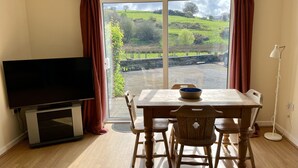A room with a TV, wooden dining table, chairs, and a view of a green landscape through a glass door. - Cottage In Dôl Y Bont, Near Aberystwyth, Ceredigion, Wales. (Dol y bont)