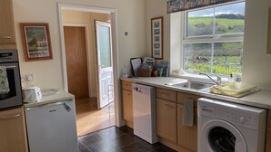 A kitchen with a washing machine, dishwasher, and oven. There is a window with a view of greenery outside. - Cottage In Dôl Y Bont, Near Aberystwyth, Ceredigion, Wales. (Dol y bont)