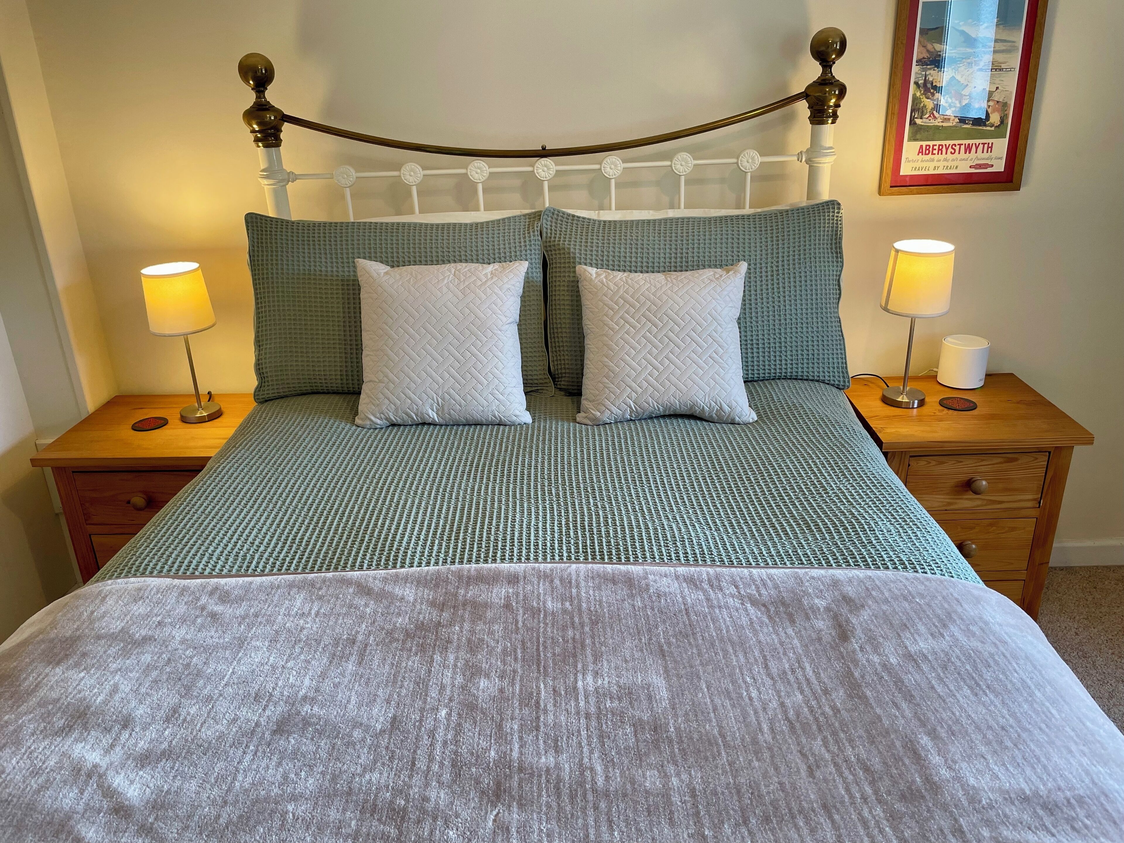 Una cama bien tendida con un edredón verde y dos almohadas, flanqueada por mesitas de noche de madera con lámparas y un cuadro enmarcado.