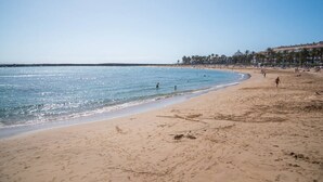 Una playa con agua azul cristalina, orilla arenosa y edificios al fondo.