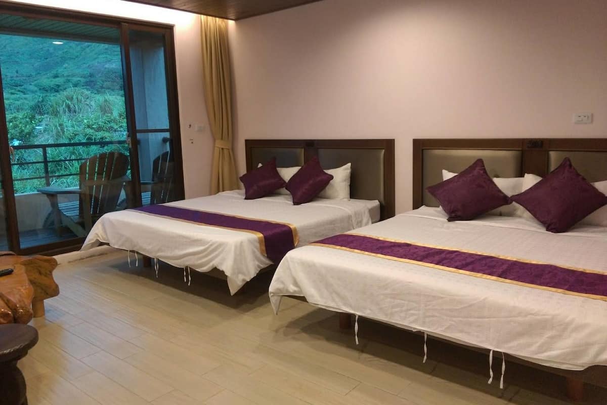 Habitación de hotel con dos camas, cada una con ropa blanca y detalles morados, piso de madera y vistas a la vegetación a través de una puerta corrediza.
