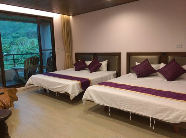 A hotel room with two beds, each with white linens and purple accents, a wooden floor, and a view of greenery through a sliding door.
