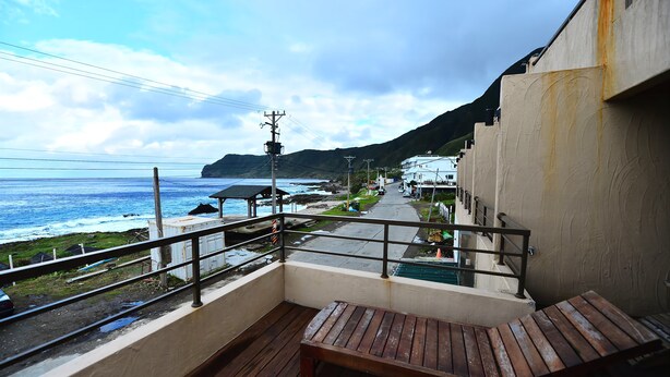 A balcony with a wooden bench overlooking a coastal area with a view of the ocean and a mountain in the background. - Two Fish (Lanyu)
