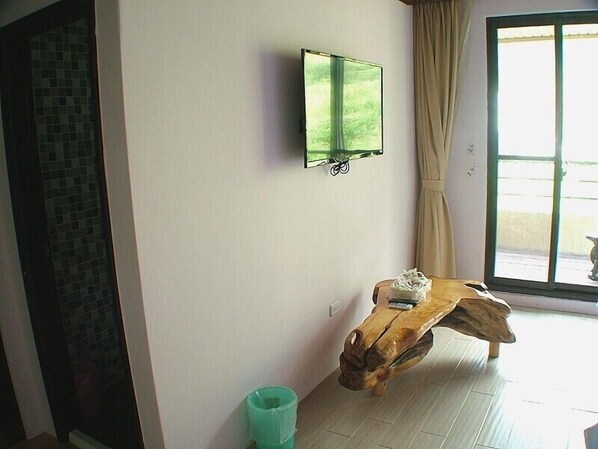 A room with a wooden coffee table, a green trash bin, a mounted TV, and a window with curtains.